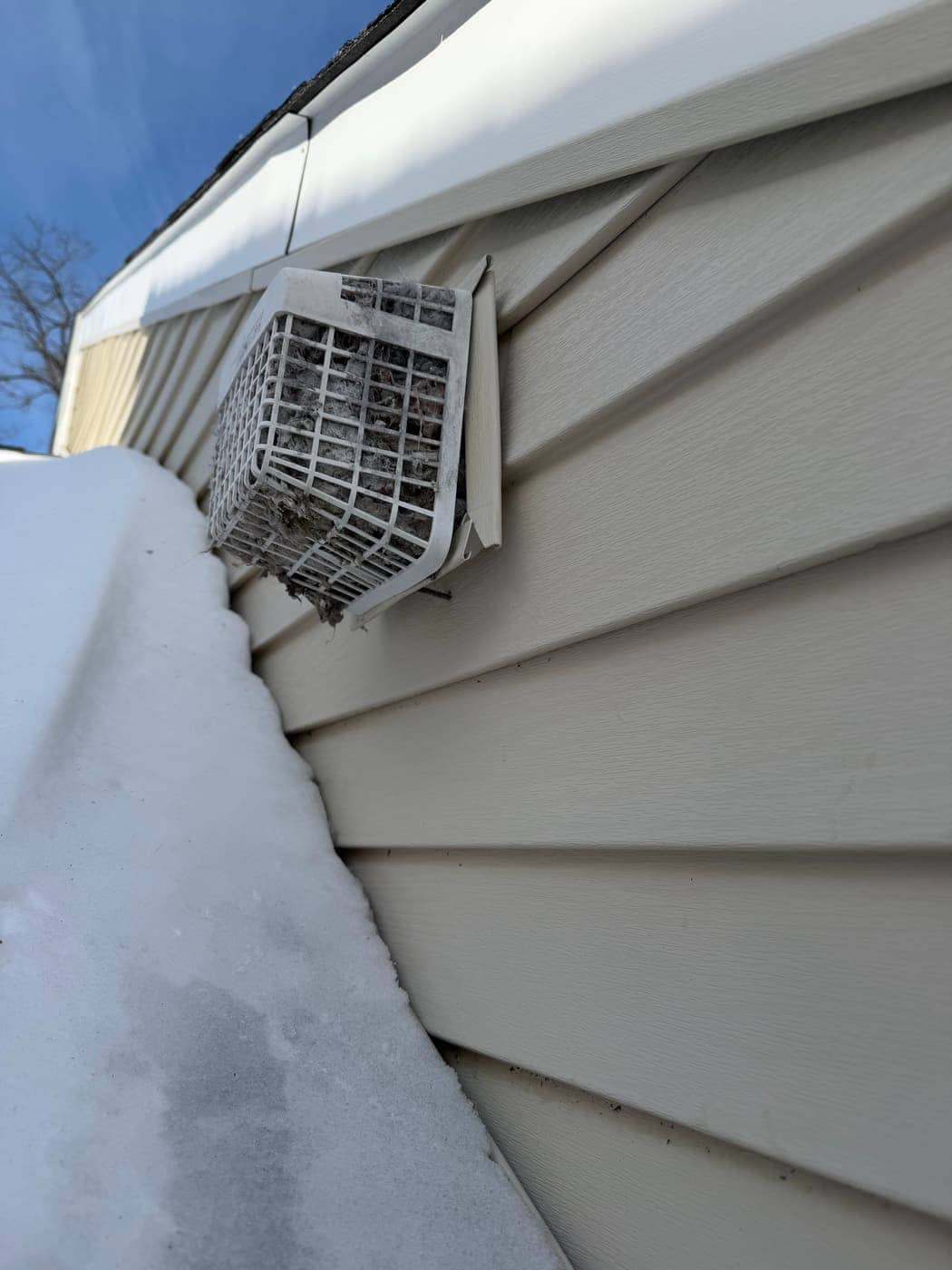 A dryer vent hood on vinyl siding with its louvers packed with lint, above a roof covered in snow.