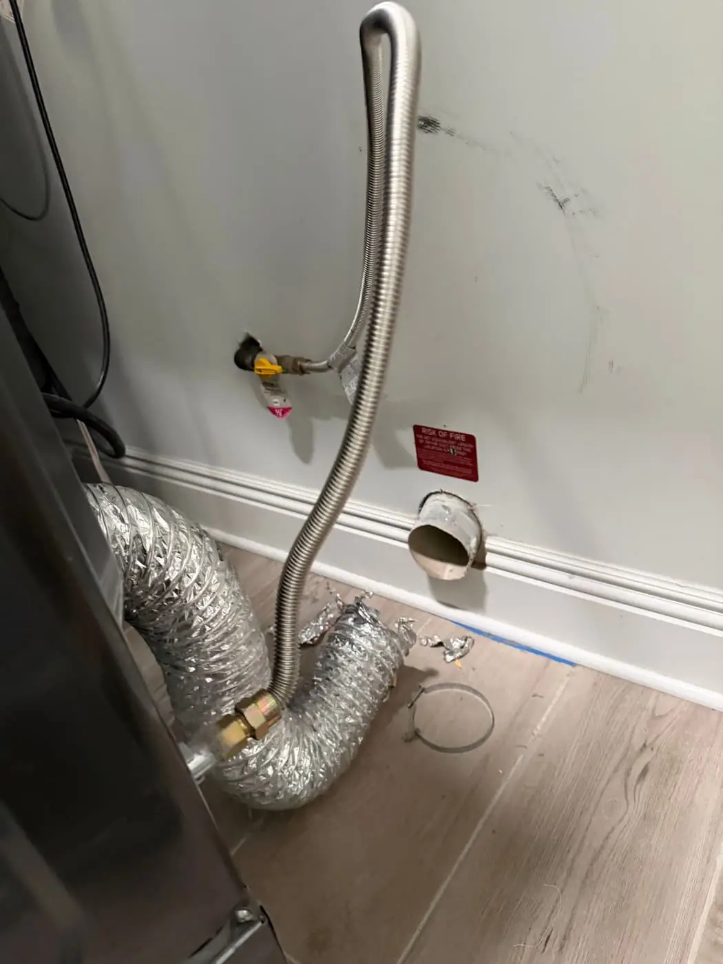 Behind a dryer: the gas line, a crumpled foil transition duct and the round vent outlet in the wall.