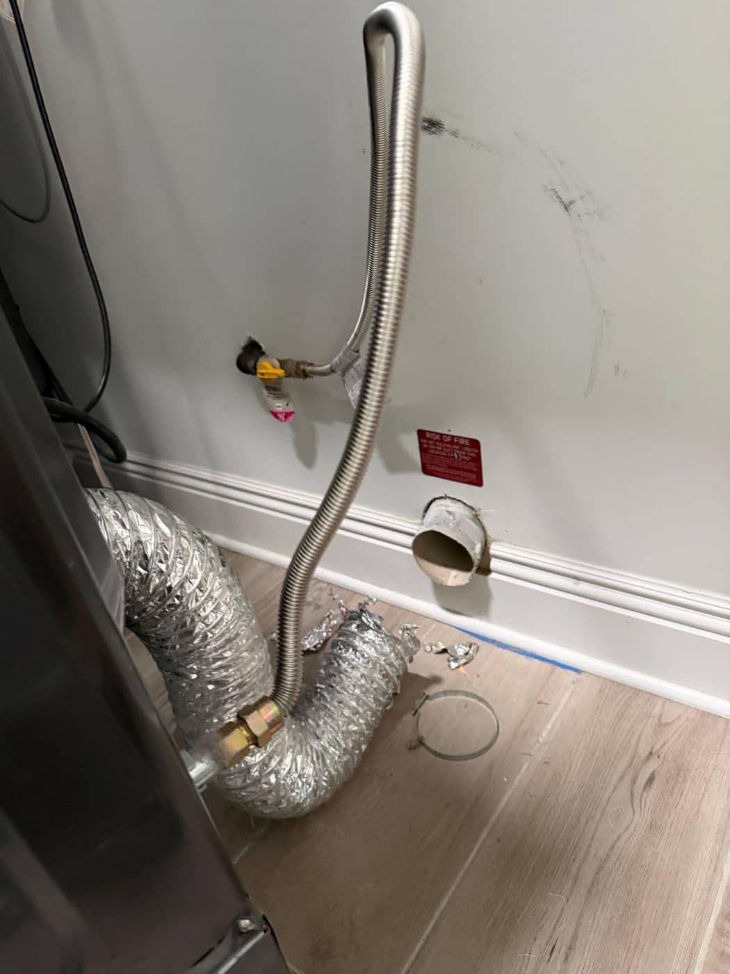 Behind a dryer: the gas line, a crumpled foil transition duct and the round vent outlet in the wall.