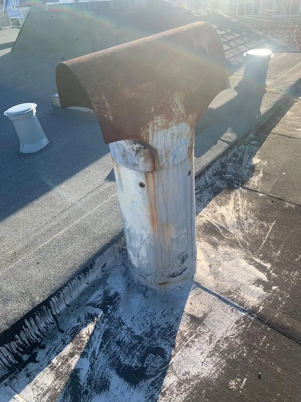 A rusted half-round cap on a metal vent pipe on a flat roof, with other roof vents behind it.