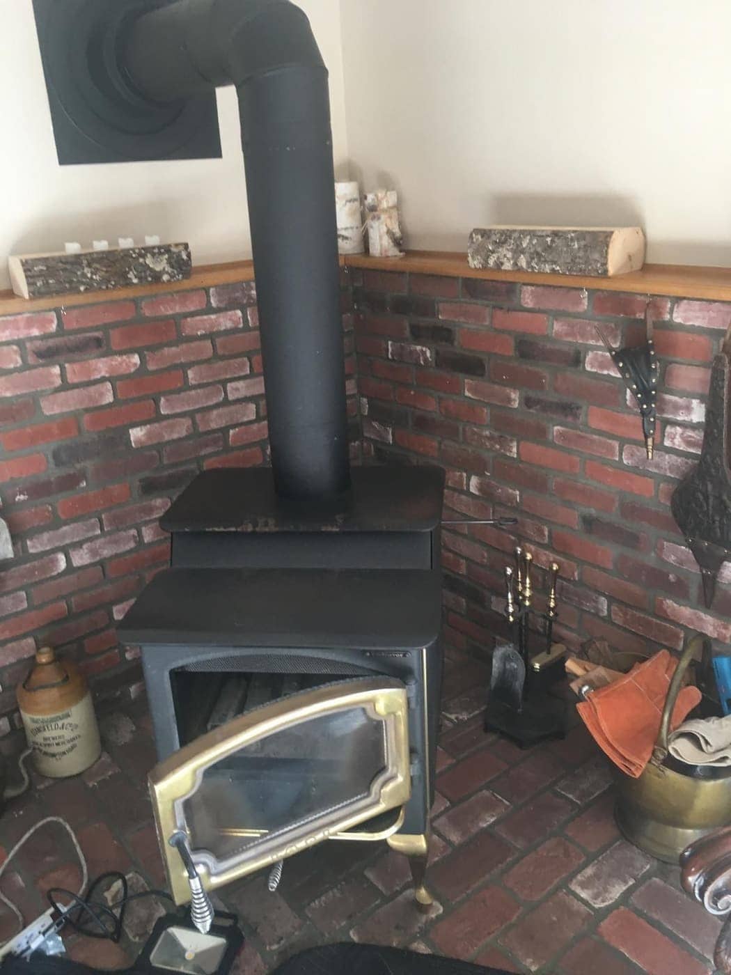 A black wood stove with a glass door and its stovepipe, set in a brick corner on a brick hearth.